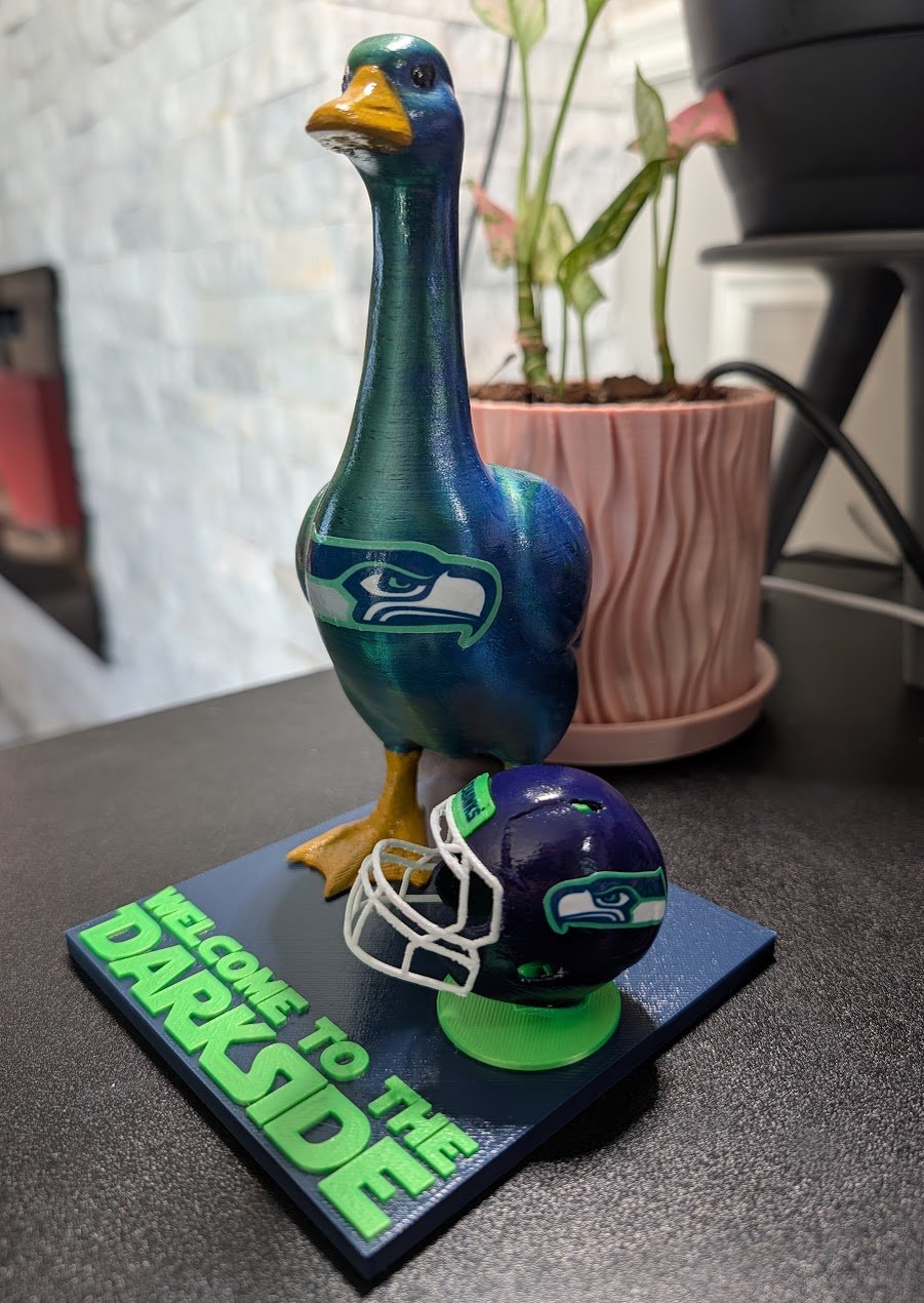 Darkside, a teal goose with a Seahawks logo, standing on a Welcome to the Darkside base next to a tiny helmet