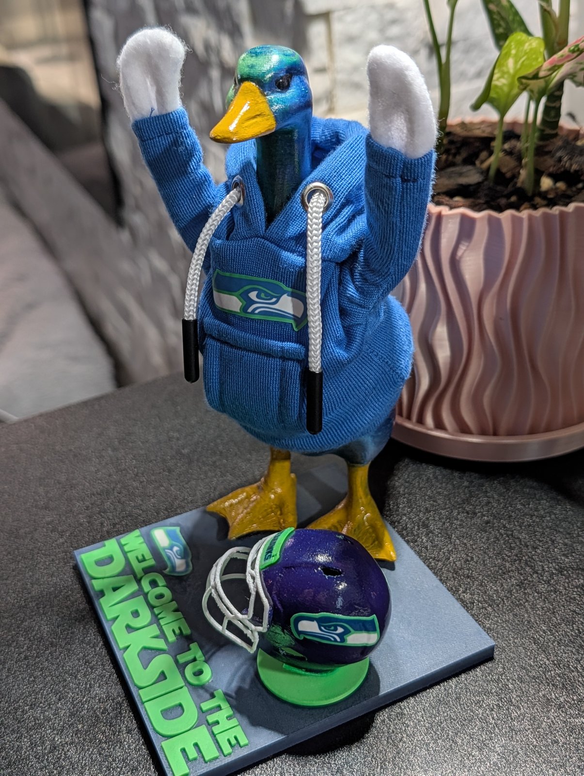 Darkside the goose wearing a blue Seahawks hoodie with both wings raised, helmet on the ground