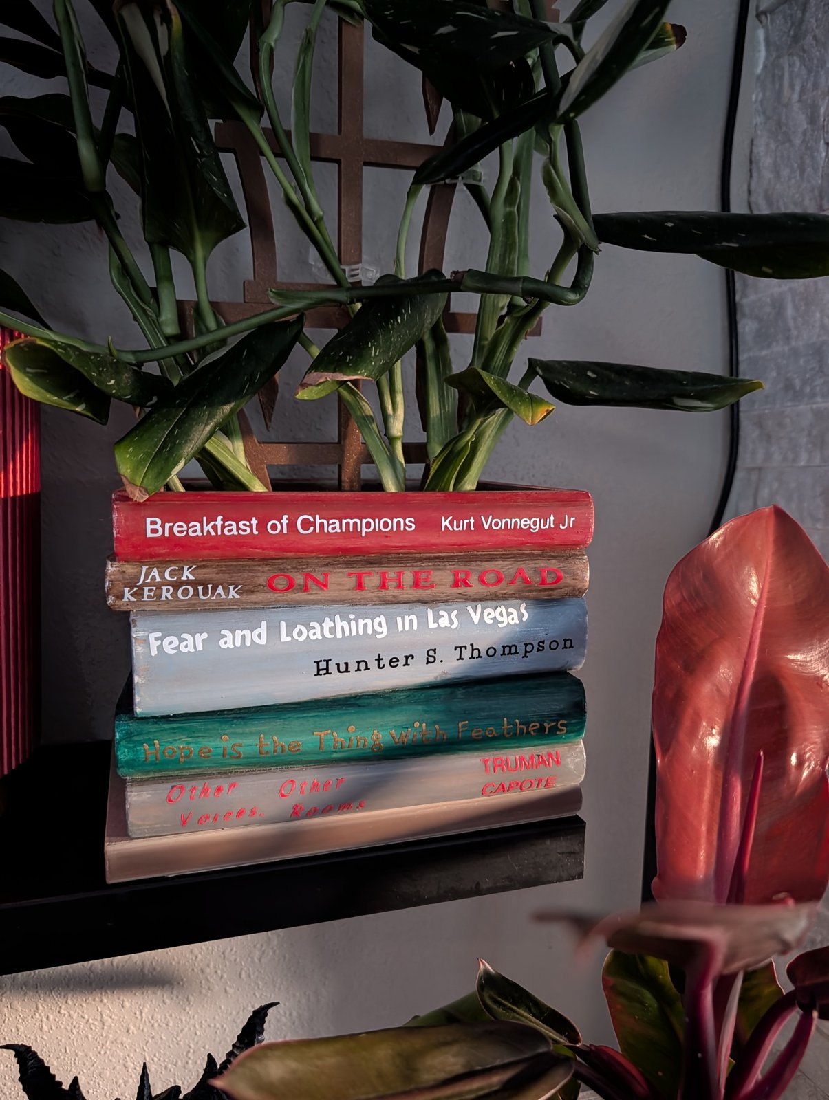 A book-stack planter on a shelf at dusk, with painted book titles like Breakfast of Champions and On the Road