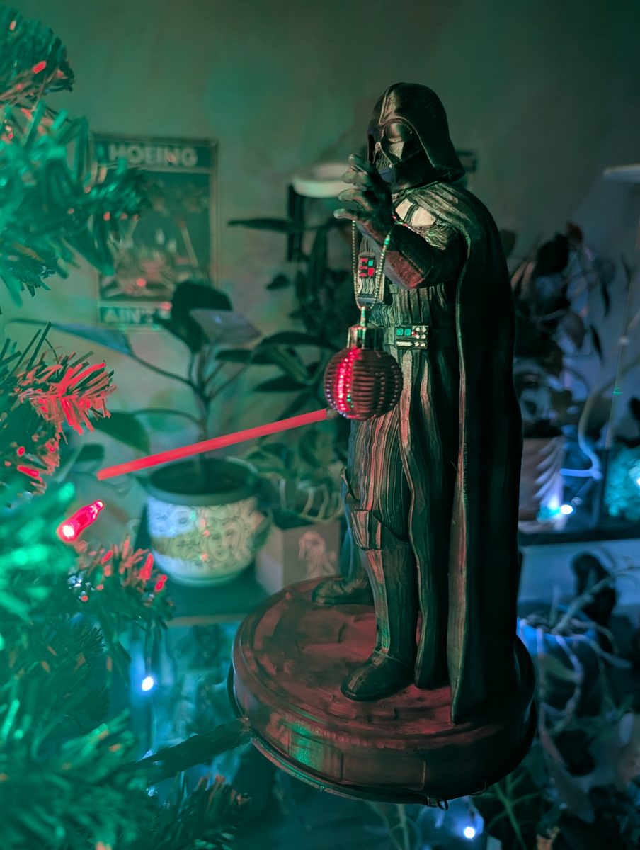 Darth Vader on a floating tree platform at night, holding a red ornament with his lit red lightsaber among Christmas lights